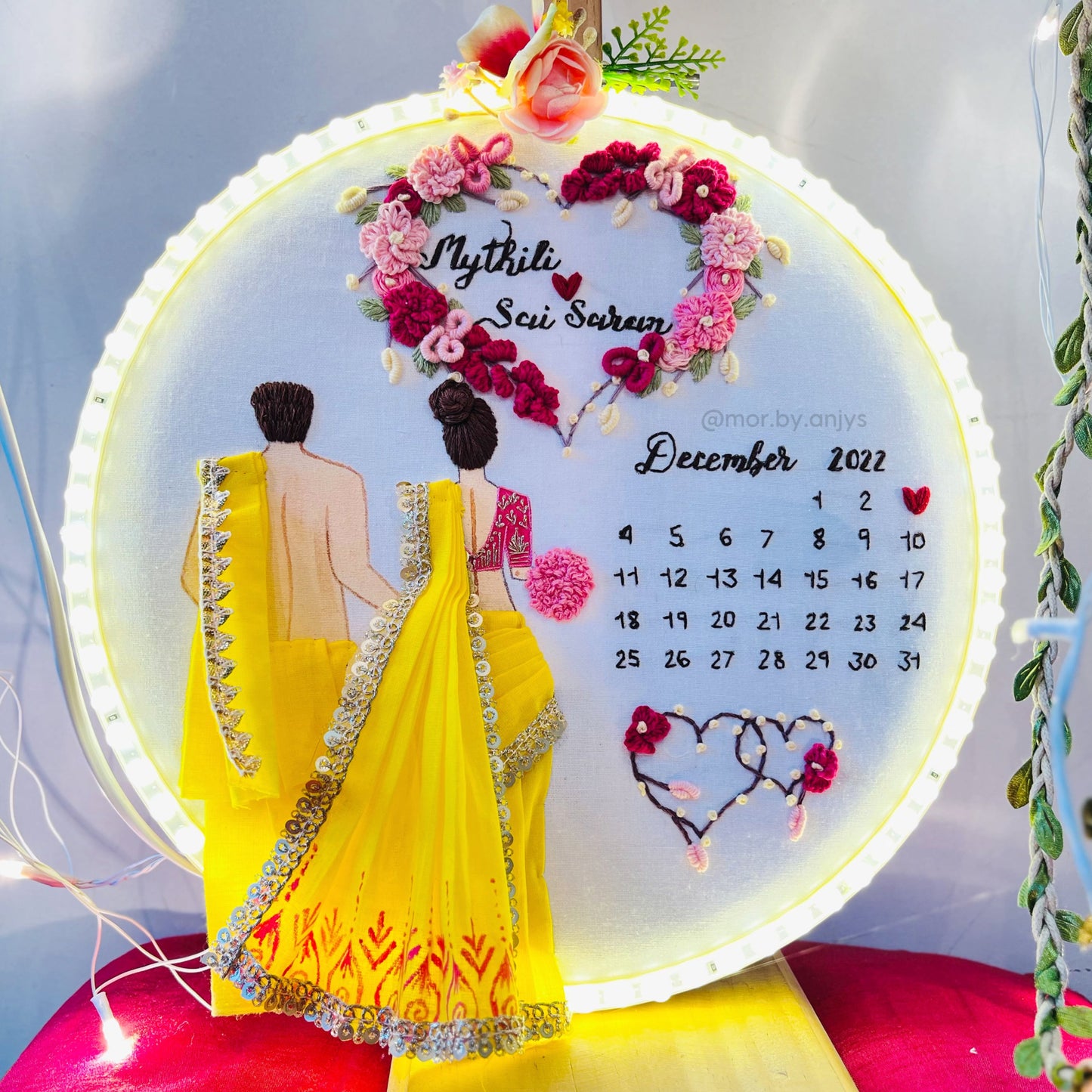 Decorative wall art with a couple in yellow sarees and floral design, featuring a calendar.