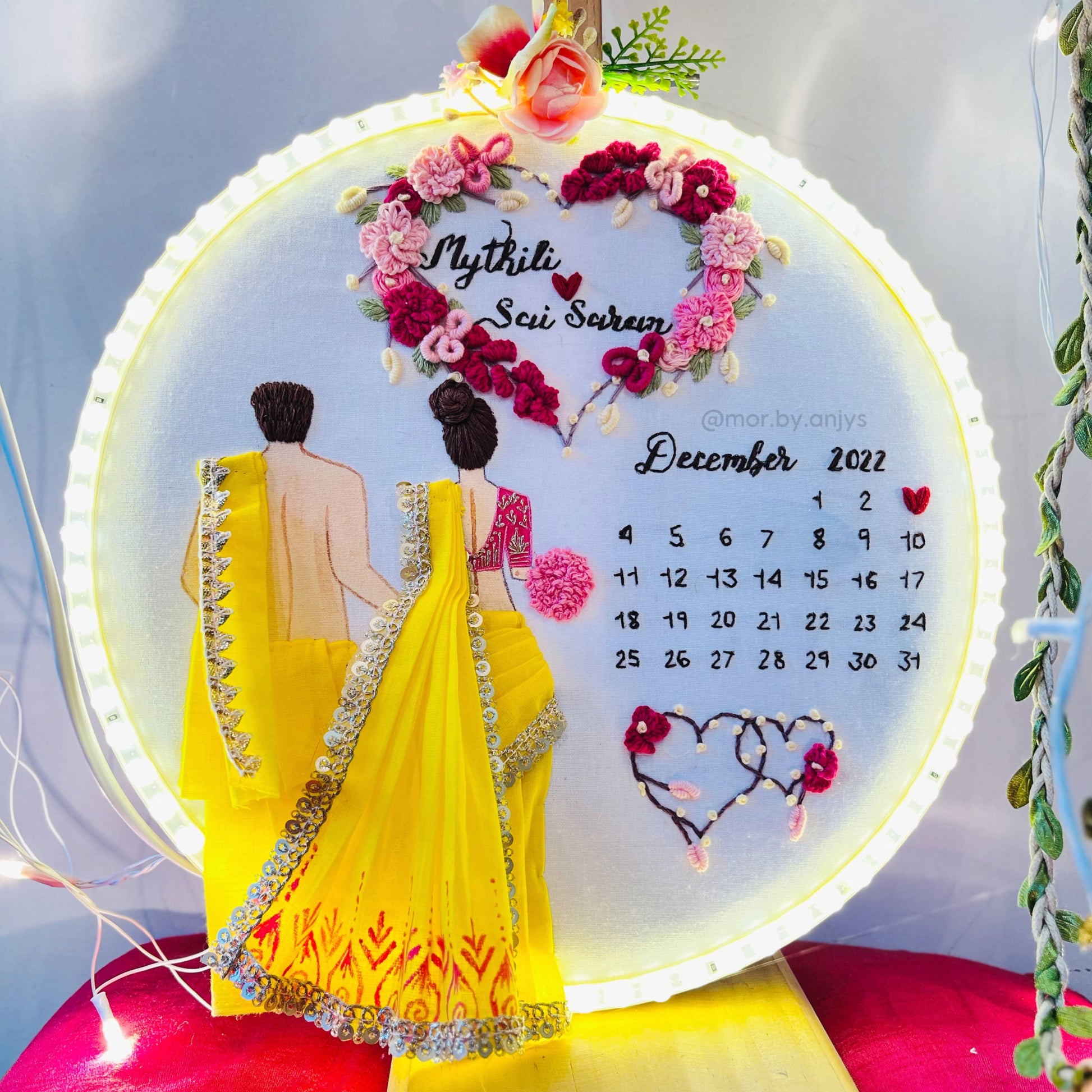 Decorative wall art with a couple in yellow sarees and floral design, featuring a calendar.