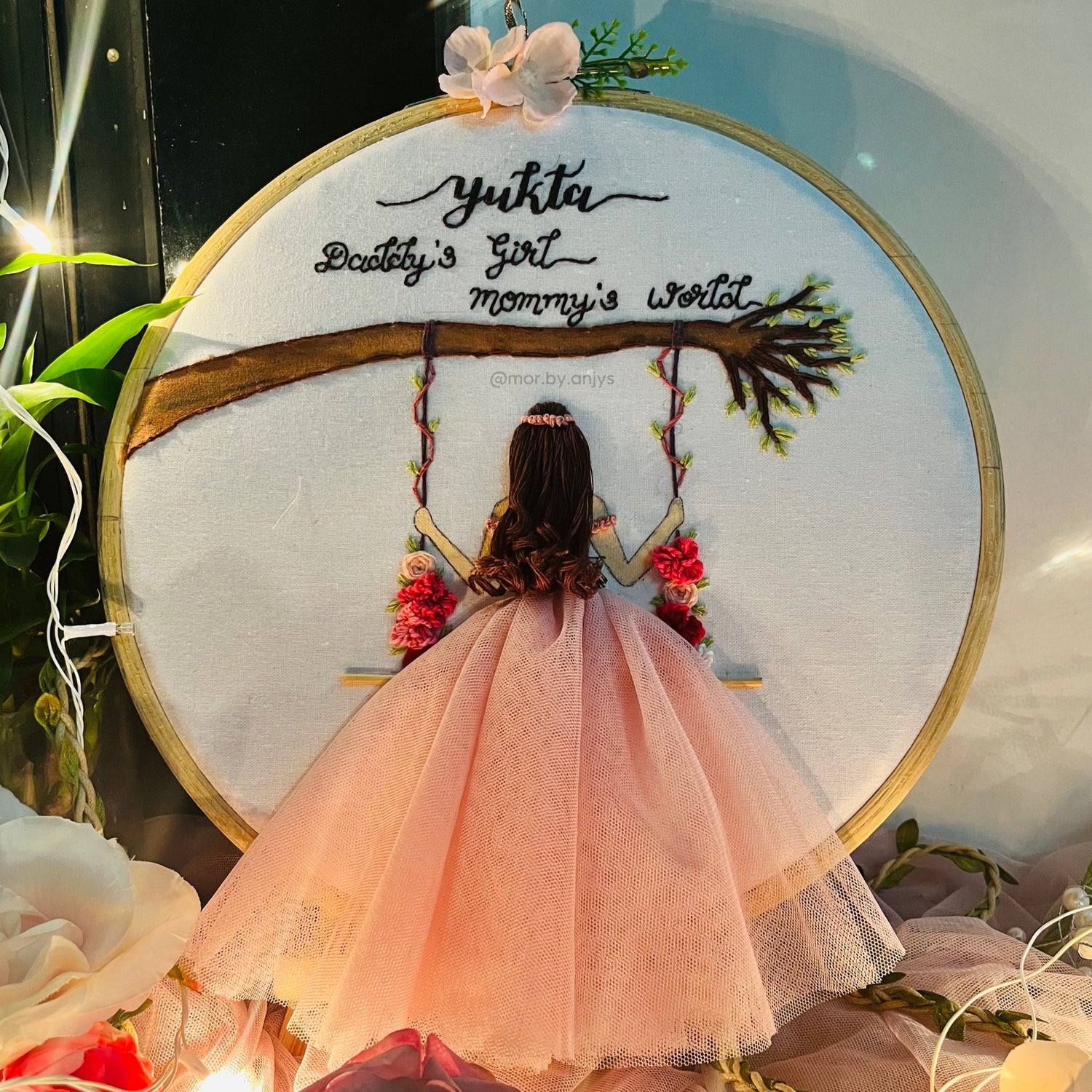 Embroidery hoop with a design of a woman on a swing, surrounded by flowers and lights.