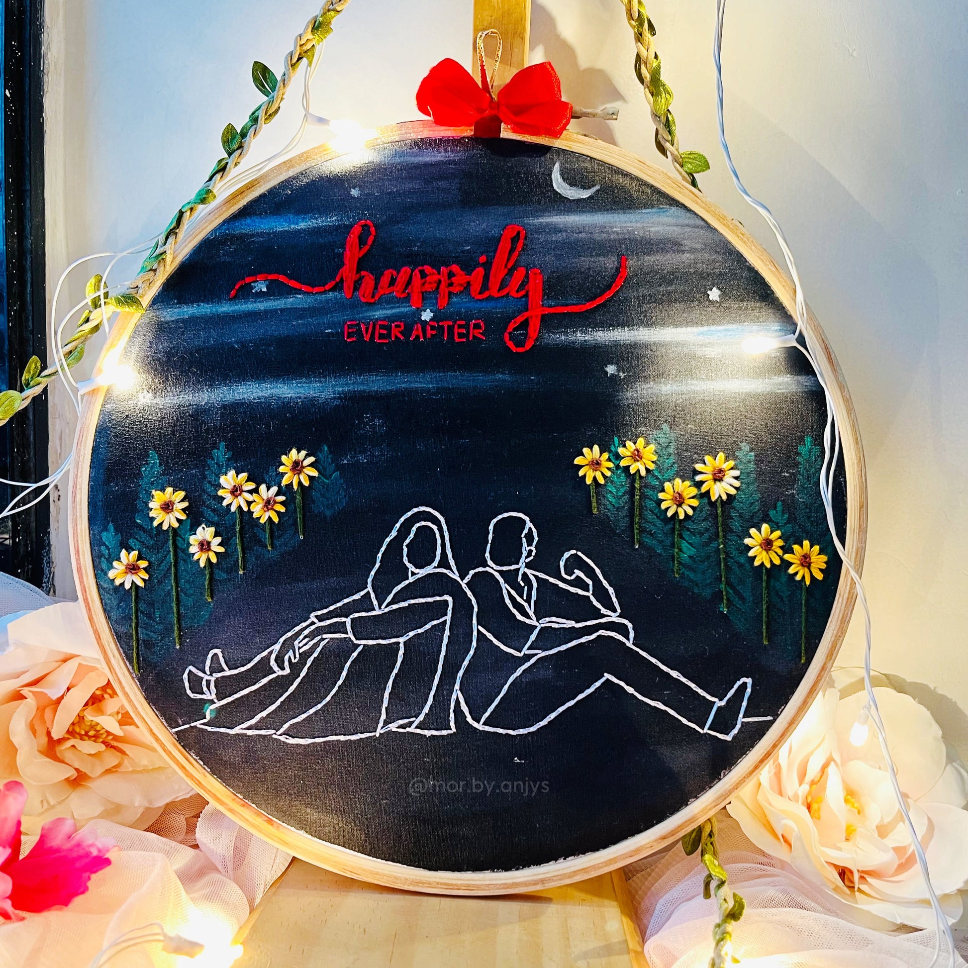 Decorative hoop with 'Happily Ever After' text, flowers, and a couple outline on a dark background.
