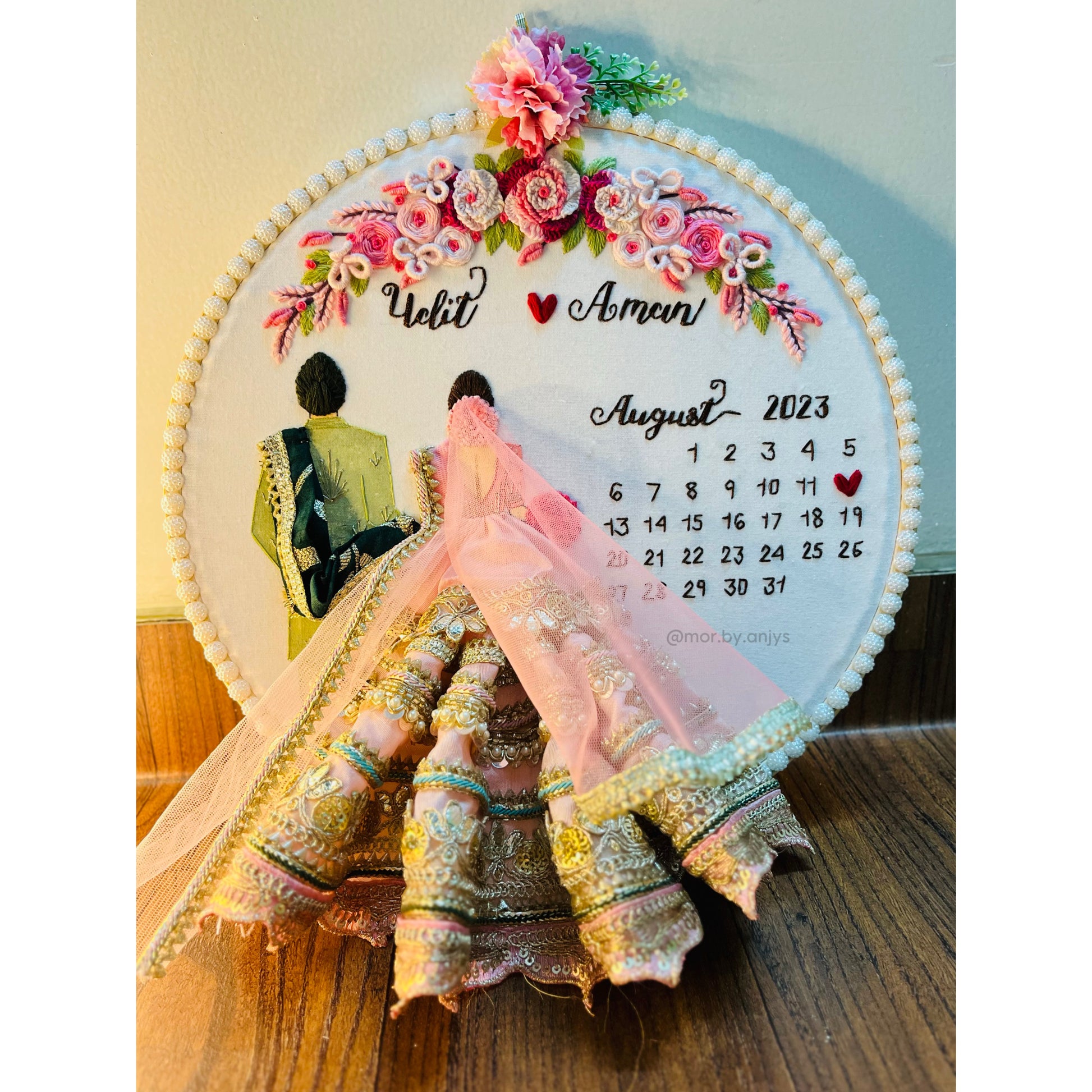 Decorative calendar with floral design and couple's names for August 2023 on a wooden surface.