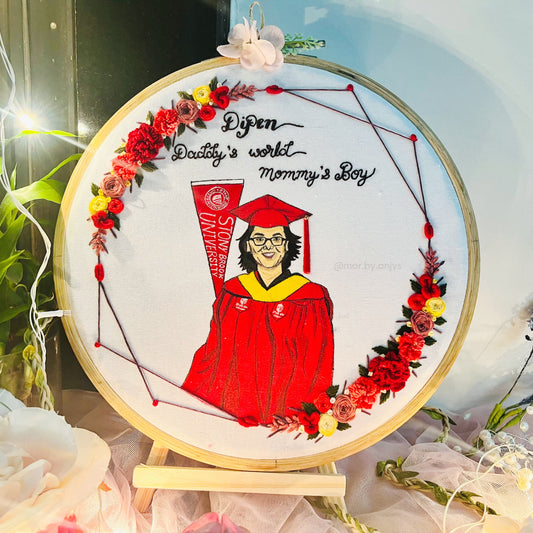 Decorative panel with a graduate in a red gown and cap, surrounded by flowers and text, on a stand.